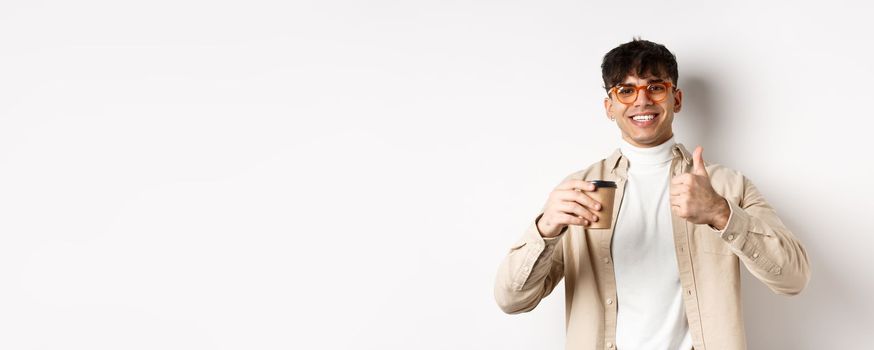 Real people. Cheerful smiling guy in glasses drinking coffee and showing thumb up, recommending cafe, standing on white background.