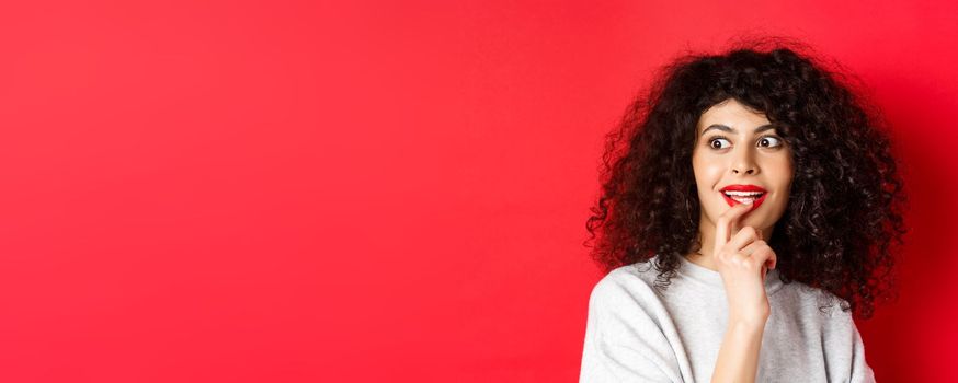 Close-up of excited curly woman touching lips and looking aside with interest, checking out cool promo, standing against red background.