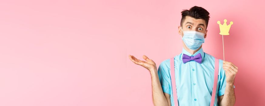 Covid, pandemic and quarantine concept. Clueless and confused young man in medical mask shrugging, showing little party crown, know nothing, standing on pink background.