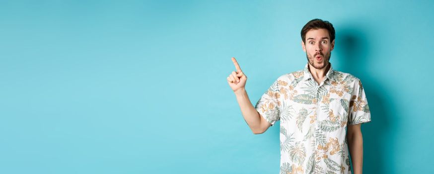 Surprised and amazed tourist in hawaiian shirt pointing finger left, showing promo and staring at camera, standing on blue background.