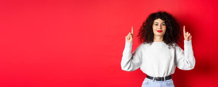 Image of cheerful smiling woman with curly hairstyle and red lips, pointing fingers up at empty space, showing advertisement, standing in casual clothes on studio background.