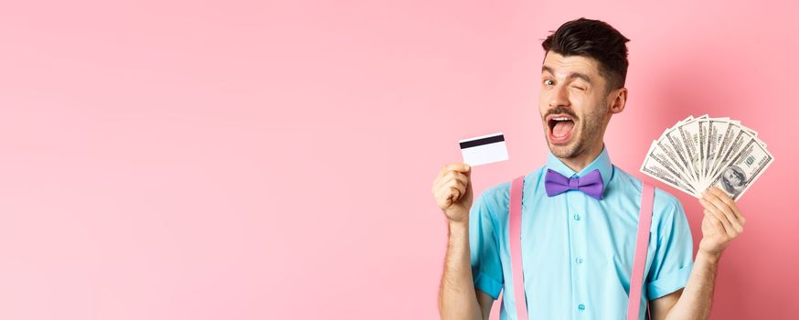 Cheerful caucasian man in bow-tie winking at you, showing plastic credit card and money, recommending special deal, standing on pink background.