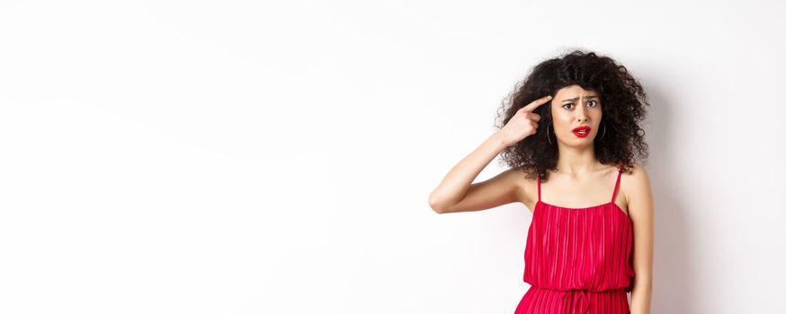 Shocked and confused caucasian woman in red dress scolding someone crazy, pointing finger at head and frowning, standing over white background.