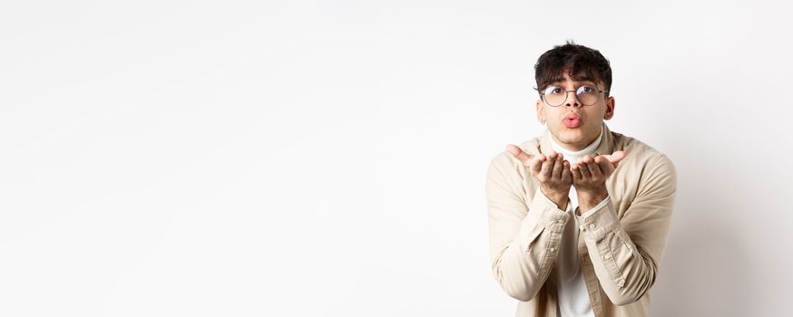 Cute boyfriend sending air kiss at camera on happy Valentines day, looking tender at lover, standing in casual clothes and glasses on white background.