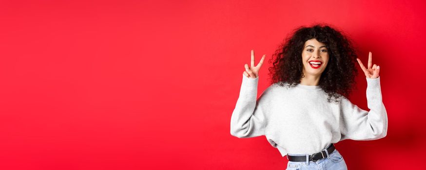 Stylish young woman with curly hairstyle, smiling happy and showing peace signs, standing in casual sweatshirt on red background.