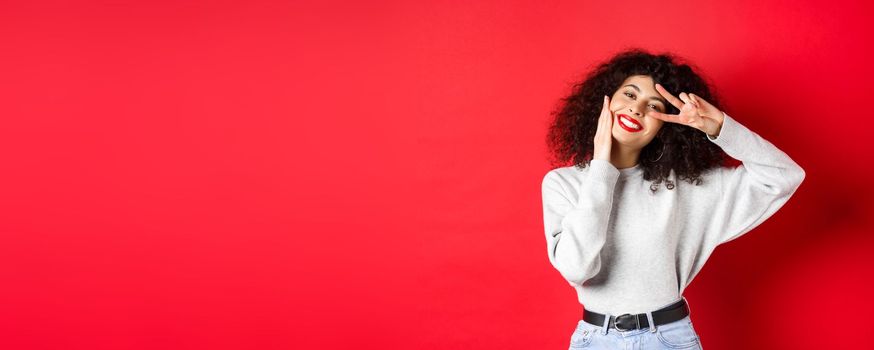 Beauty and makeup. Happy young woman with curly hair, touching face and showing v-sign with cute smile, standing against red background.