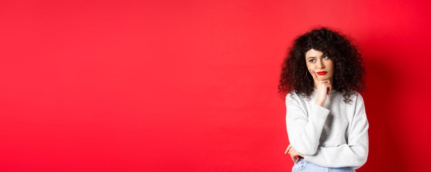 Grumpy young woman with curly hair, looking annoyed or bored at empty space, standing pensive and sad on red background.