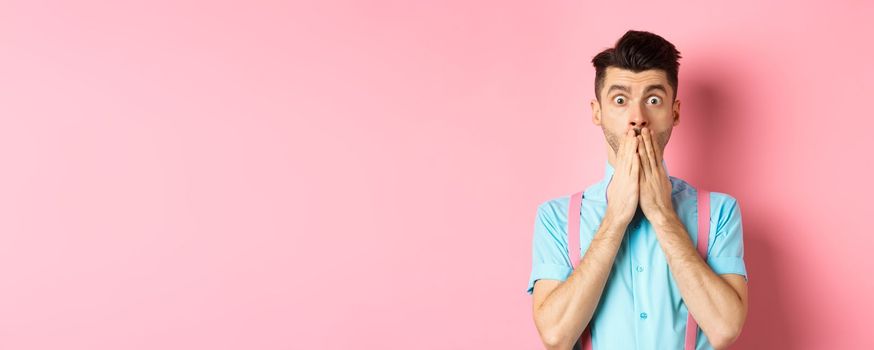 Shocked man looking in startled, covering mouth with hands and staring at camera, standing on pink background.