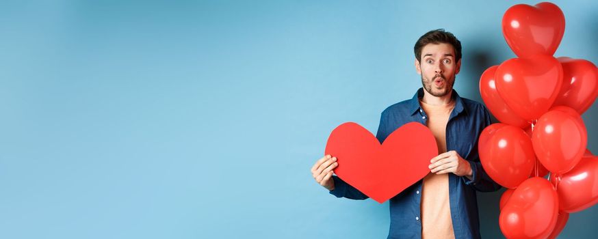 Valentines day concept. Boyfriend in love looking amazed at camera, holding red paper heart and standing with romantic balloon, blue background.