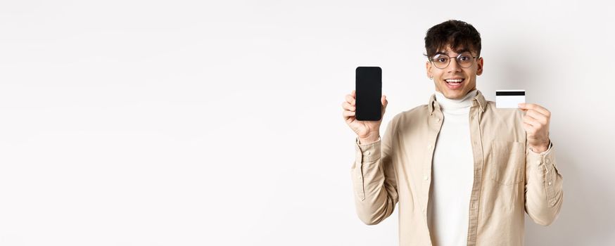 Online shopping. Surprised and happy young man showing credit card and mobile phone screen, standing on white background.