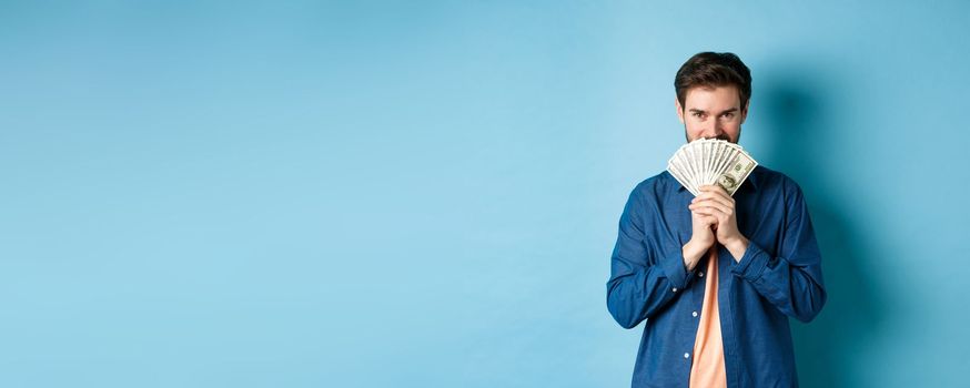 Young man hiding smiling face behind money and looking determined at camera, standing on blue background.