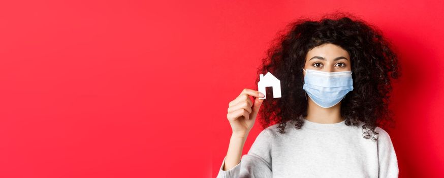 Real estate and covid-19 concept. Excited woman in medical mask showing small paper house cutout, standing on red background.