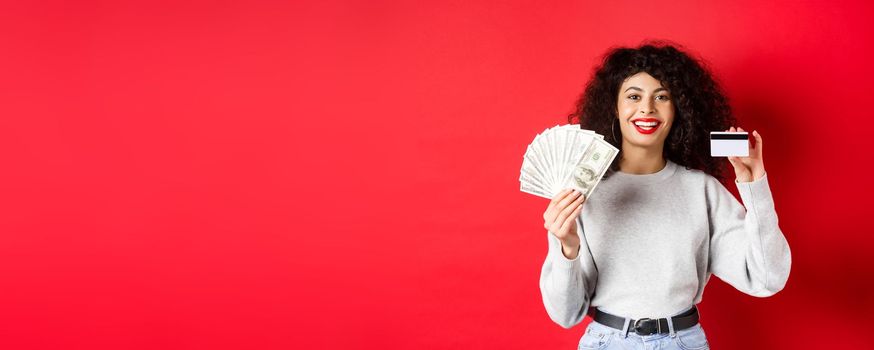Portrait of stylish young woman with curly hair, showing money in cash and plastic credit card, red background.