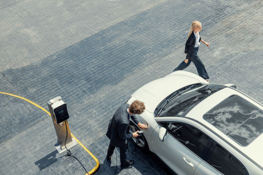 Aerial view progressive lifestyle of businesspeople with electric car connected to charging station at city center public parking car. Eco friendly rechargeable car powered by alternative clean energy