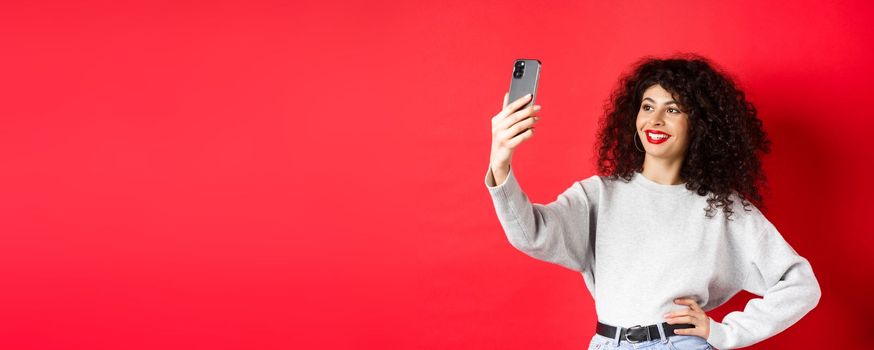 Image of stylish female blogger taking selfie on smartphone, posing for photo on mobile phone, standing at red background.