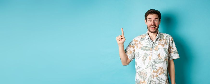 Excited guy in hawaiian shirt talking about vacation, looking amazed and pointing up, standing on blue background.
