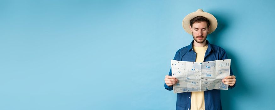 Handsome tourist with summer hat reading map, looking for sightseeing on vacation, standing on blue background.