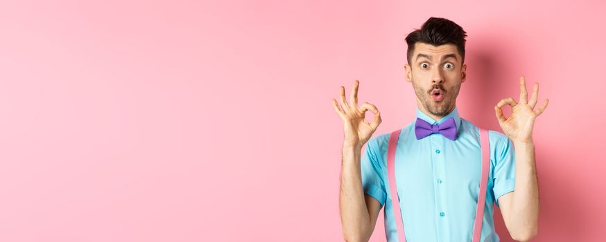Impressed caucasian guy in bow tie, checking out cool offer, showing okay gesture and saying wow, praising good product, standing on pink background.