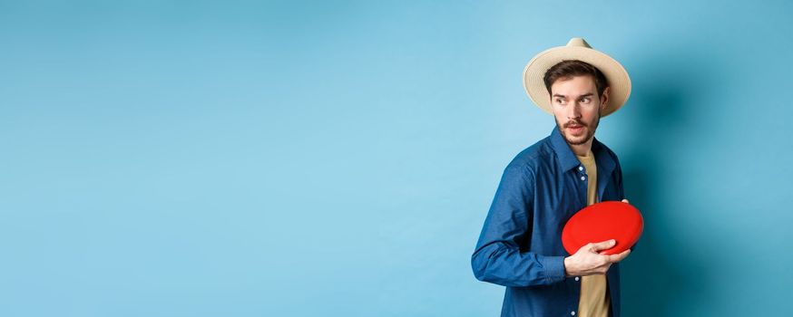 Handsome young guy in summer hat playing frisbee, throwing it aside, standing on blue background.