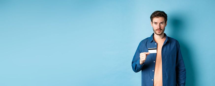 Handsome young man with beard, wearing casual clothes, showing plastic credit card and looking at camera, blue background.