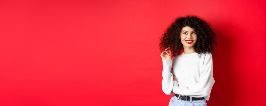 Modern woman in sweatshirt, playing with curly hair and looking aside at empty space, standing on red background.