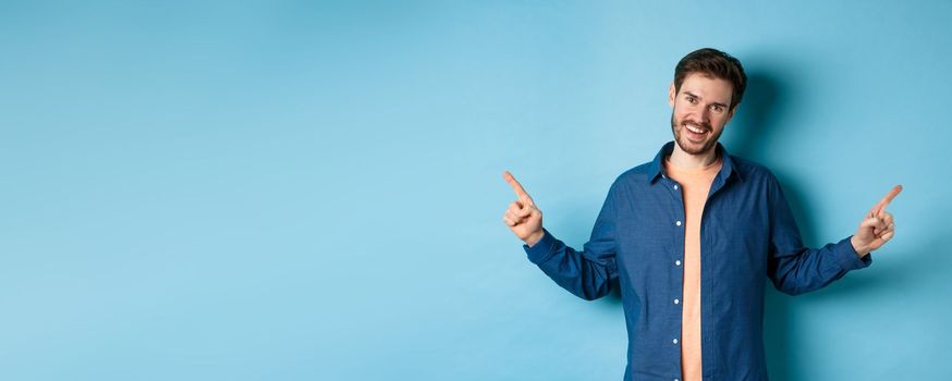 Handsome caucasian man with beard, smiling with white teeth and pointing fingers sideways, showing two choices, standing on blue background.