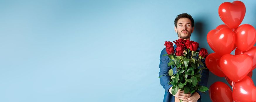 Romantic boyfriend confessing in love and giving bouquet of roses, pucker lips for kiss, standing with heart balloons on blue background.