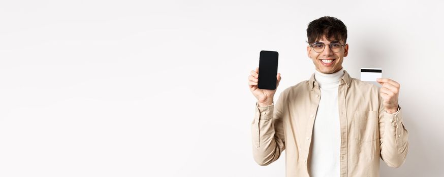 Online shopping. Natural guy in glasses showing empty smartphone screen and plastic credit card, smiling pleased, recommending bank, standing on white background.