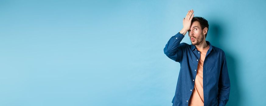 Annoyed man making facepalm and roll eyes from something stupid, standing over blue background with hand on forehead.