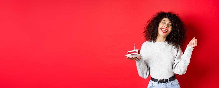 Celebration and holidays concept. Happy beautiful woman dancing and making birthday wish, holding b-day cake and smiling, standing on red background.