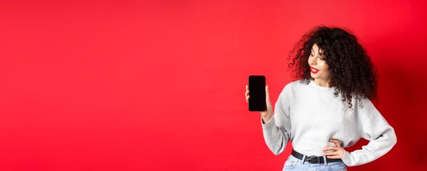 Portrait of stylish young woman with curly hair showing empty smartphone screen, demonstrating shopping app, standing on red background.