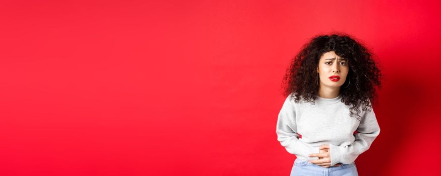 Image of young woman having stomach ache, bending from pain and complaining on painful menstrual cramps, standing on red background.