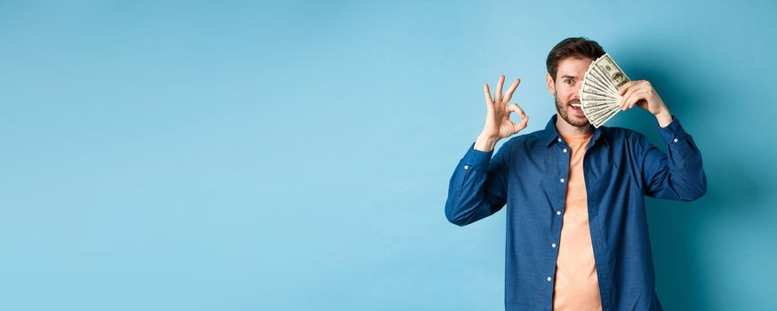 Cheerful young man showing OK sign and cover half of face with dollars, standing on blue background.