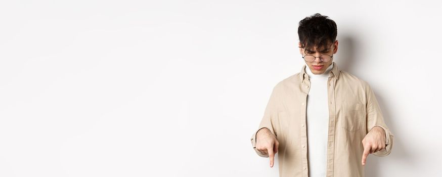 Confused frowning guy in glasses looking, pointing fingers down at something strange, standing puzzled on white background.
