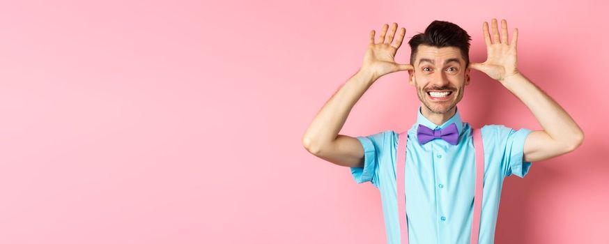 Silly and childish guy in suspenders and bow-tie, mocking someone, fooling around and making funny gestures, standing on pink background.