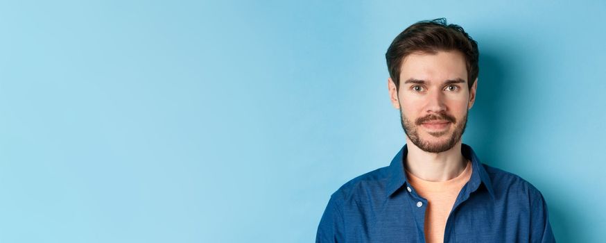 Close-up of young caucasian guy with beard smiling at looking happy at camera, standing on blue background.