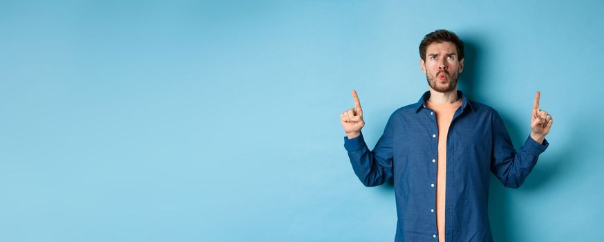 Confused young man frowning, pointing fingers up and looking displeased, standing on blue background. Copy space
