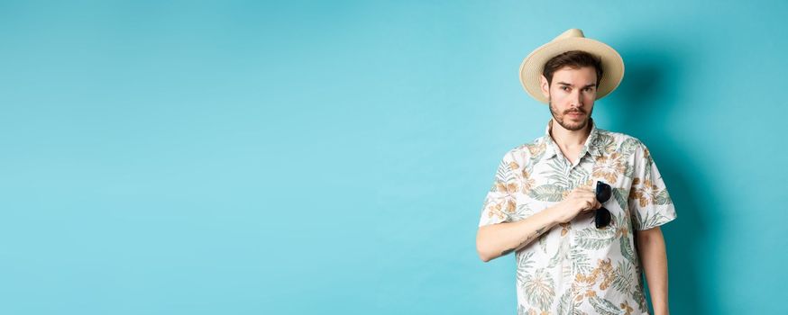 Handsome tourist in hawaiian shirt and summer hat, put sunglasses in pocket, going on vacation, standing against blue background.