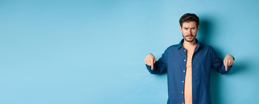 Concerned young man, frowning and looking worried, pointing fingers down, showing something bad, standing on blue background.