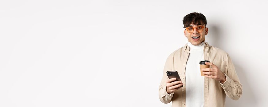 Cheerful smiling young man in glasses drinking coffee and holding smartphone, looking excited at camera, standing on white background.