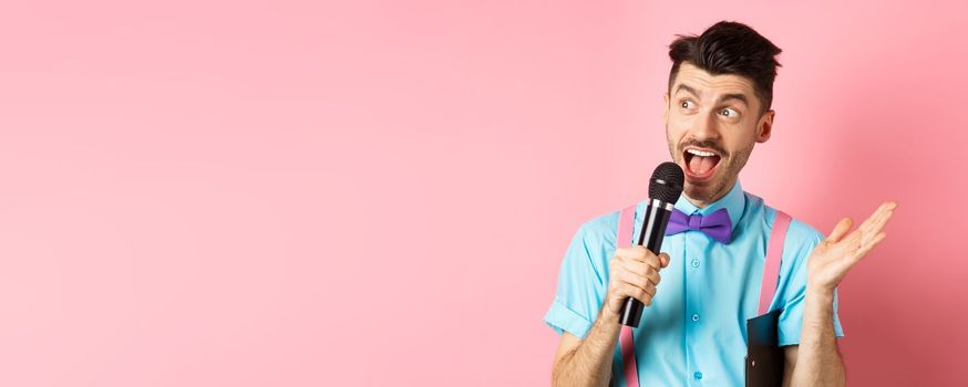 Image of male show host making speech, talking in microphone with clipboard under armpit, enteratin people on festive event, standing over pink background.