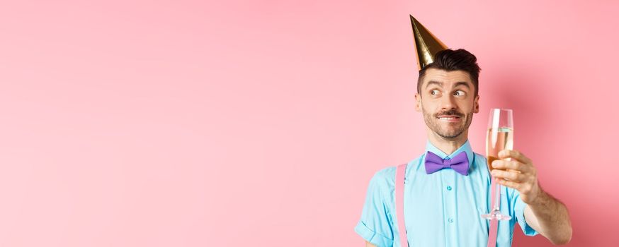 Holidays and celebration concept. Cheerful young man celebrating birthday in party hat, thinking of speech, raising glass of chamapgne for toast, pink background.