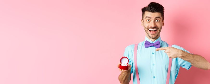 Valentines day. Cheerful young man going to make proposal, pointing finger at engagement ring and smiling excited, standing on romantic pink background.