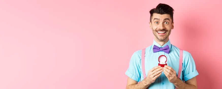Valentines day. Romantic man in bow-tie showing engagement ring and smiling, asking to marry him, making proposal to lover on pink background.