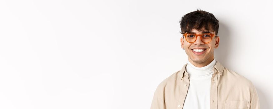 Close-up of handsome hipster guy with natural smile, wearing glasses and earrings, looking happy at camera, standing on white background.