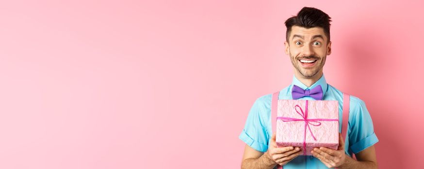 Holidays and celebration concept. Hopeful smiling guy looking with love, presenting cute gift, standing over pink background.