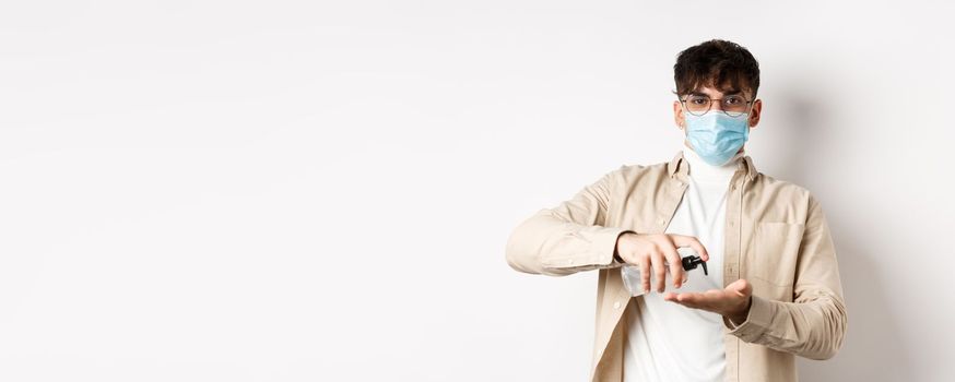 Health, covid and quarantine concept. Young hispanic guy in glasses and face mask using hand sanitizer, apply antiseptic and look at camera, standing on white background.