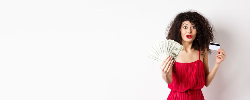 Excited woman looking with amazement and disbelief, showing dollars prize and plastic credit card, standing in red dress on white background.