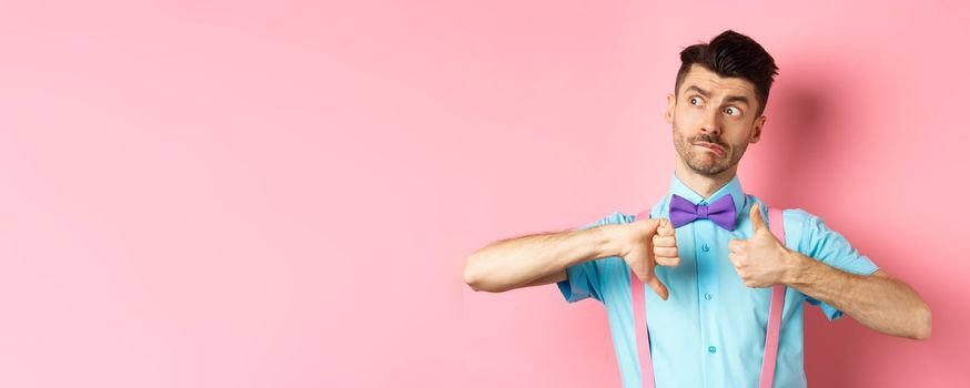 Confused guy looking pensive while judging, showing thumbs up and down, standing indecisive and unsure against pink background.