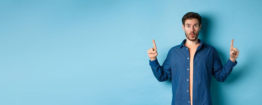 Surprised young man say wow, checking out advertisement, pointing fingers up and looking impressed, standing on blue background.